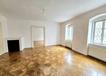 Geräumiger Raum mit Fischgrätparkett, dekorativem Kamin und hohen Fenstern, ideal für ein Wohnzimmer.