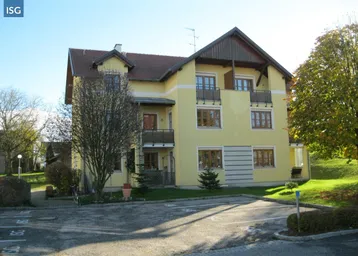 Gelbes Mehrfamilienhaus mit Satteldach, mehreren Balkonen und Stellplätzen im Freien, umgeben von Grünflächen.