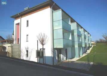Modernes Mehrfamilienhaus mit weißer Fassade, großen Fenstern und Glasbalkonen in grüner Umgebung.