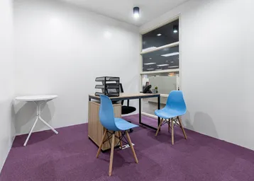 Modernes Büro mit lila Teppichboden, einem Schreibtisch und zwei blauen Stühlen. Große Fenster sorgen für natürliches Licht.