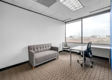 Kleines Büro mit einem Sofa, Schreibtisch und großen Fenstern, die viel Tageslicht hereinlassen.