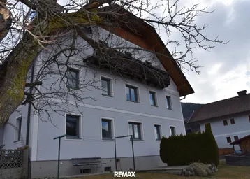 Traditionelles Wohnhaus mit heller Fassade, umgeben von einem kleinen Vorgarten und altem Baumbestand.