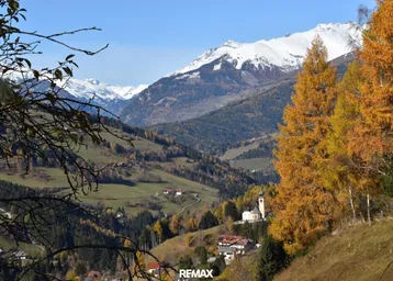 Panoramablick auf schneebedeckte Berge und ein malerisches Tal mit Herbstbäumen und einer Kirche.