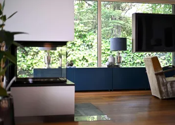 Helles Wohnzimmer mit modernem Kamin, blauen Möbeln und großen Fenstern mit Blick ins Grüne.