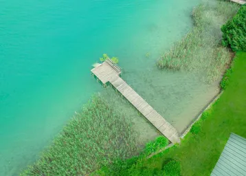 Luftaufnahme eines Holzstegs, der in einen türkisfarbenen See mit Schilf und grünen Ufern ragt.