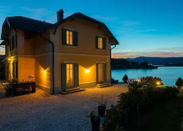 Elegantes Haus am Seeufer bei Sonnenuntergang mit beleuchteter Fassade und Garten.