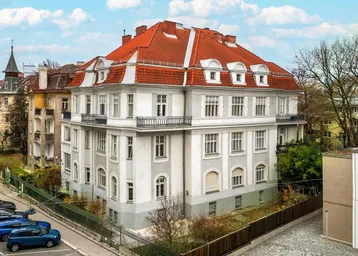 Elegantes Mehrfamilienhaus mit klassischer Architektur und rotem Ziegeldach in städtischer Umgebung.