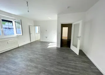 Helles, leeres Wohnzimmer mit modernem Vinylboden und Blick ins Grüne durch die Fenster.
