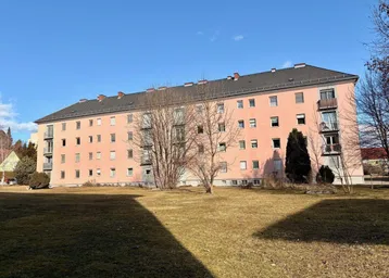 Großes rosafarbenes Mehrfamilienhaus mit vielen Fenstern und Balkonen, umgeben von einer Grünfläche.