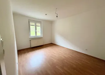 Helles Zimmer mit Laminatboden, weißen Wänden und einem Fenster mit Rollläden.