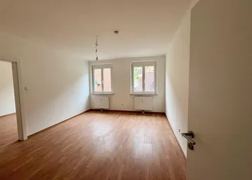 Helles, leeres Zimmer mit Laminatboden und zwei Fenstern, bereit zur individuellen Gestaltung.