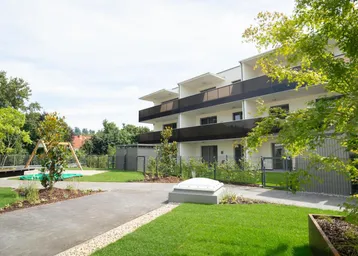 Moderner Apartmentkomplex mit Gemeinschaftsgarten, Spielplatz und Balkonen unter blauem Himmel.
