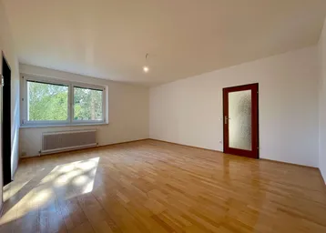 Helles, leeres Zimmer mit Parkettboden und einem Fenster, das viel Tageslicht hereinlässt.