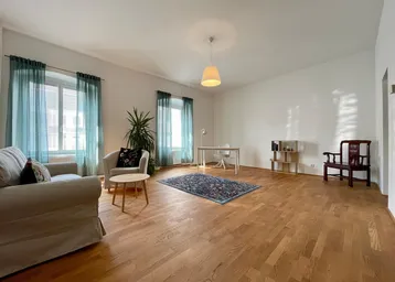 Geräumiges Wohnzimmer mit Holzboden, zwei Fenstern und einem Arbeitsbereich im Hintergrund.