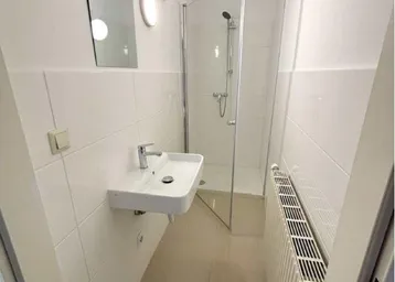 Schlichtes Badezimmer mit Duschkabine, Waschbecken und Heizkörper, hell gefliest.