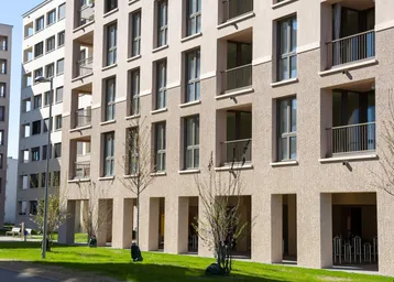 Moderne Wohngebäude-Fassade mit Balkonen, großen Fenstern und begrüntem Außenbereich im Sonnenlicht.
