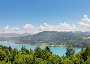 Panoramablick auf einen See umgeben von bewaldeten Hügeln und fernen Bergen unter blauem Himmel.