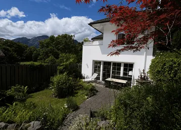 Weißes Haus mit sonniger Terrasse, gepflegtem Garten und beeindruckendem Bergblick unter blauem Himmel.