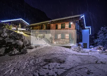 Charmantes Holzchalet in verschneiter Berglandschaft bei Nacht, mit beleuchteter Fassade und Zufahrt.