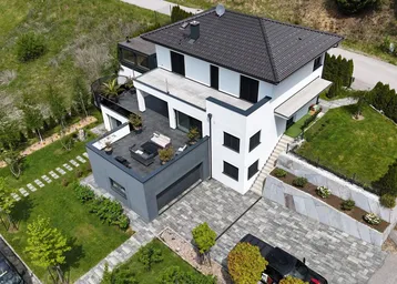 Moderne Villa mit dunklem Dach, weißer Fassade, mehreren Terrassen und gepflegtem Gartenbereich.