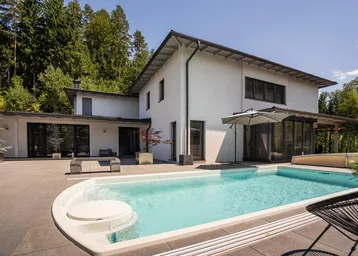 Modernes Haus mit großem Pool und weitläufiger Terrasse, umgeben von grünen Bäumen unter blauem Himmel.