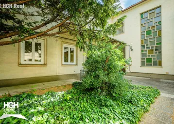 Außenansicht des Gebäudes mit dekorativen Fenstern, viel Grün und einem alten Baum im Vordergrund.