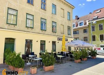Außenansicht des Gebäudes mit einer belebten Restaurantterrasse und Sonnenschirmen an einem sonnigen Tag.