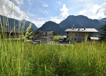 Ländliche Umgebung mit Häusern, umgeben von grünen Wiesen und Bergen unter blauem Himmel.