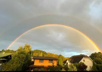 Spektakulärer doppelter Regenbogen über Wohnhäusern und bewaldeten Hügeln bei Sonnenuntergang.
