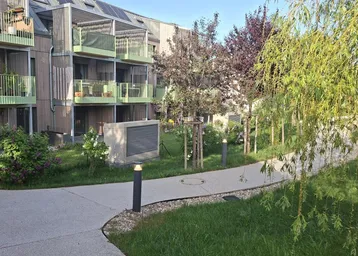 Modernes Mehrfamilienhaus mit Balkonen, begrüntem Gartenbereich, Gehwegen und Photovoltaikanlage auf dem Dach.