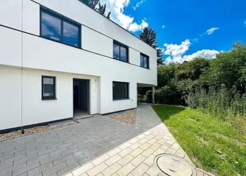 Moderne weiße Fassade eines Neubaus mit dunklen Fenstern und gepflastertem Eingangsbereich.