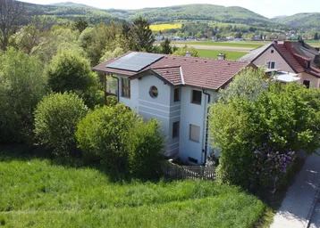 Freistehendes Haus mit Ziegeldach und Photovoltaikanlage, umgeben von viel Grün und Hügellandschaft.