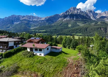 Luftaufnahme eines Hauses auf einem grünen Hügel mit majestätischer Bergkulisse im Hintergrund.