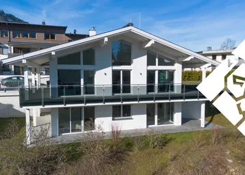 Modernes Einfamilienhaus mit großen Glasflächen, Balkon und Blick auf die umliegende Berglandschaft.