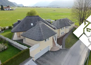 Luftaufnahme eines eleganten Hauses mit geschwungenem Dach, umgeben von weitläufigen Grünflächen und Bergen.