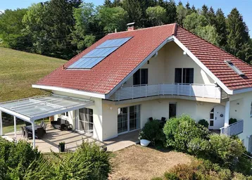Großzügiges Einfamilienhaus mit rotem Ziegeldach, Solaranlage und überdachter Terrasse in grüner Umgebung.