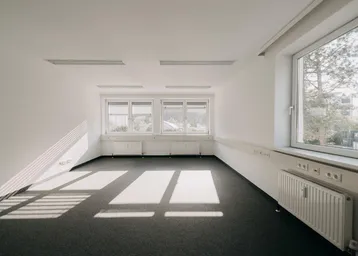 Helles, leeres Büro mit Teppichboden und großen Fenstern, die viel Tageslicht hereinlassen.