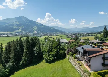 Panoramablick auf Häuser in einer grünen Talandschaft mit majestätischen Bergen im Hintergrund unter blauem Himmel.