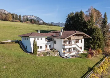 Traditionelles Haus im Alpenstil mit mehreren Balkonen und Bergblick, umgeben von grünen Wiesen und Bäumen.