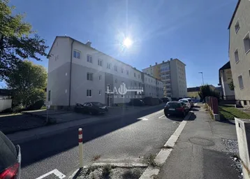 Mehrstöckiges Wohngebäude mit heller Fassade und Parkplätzen an der Straße unter strahlendem Sonnenschein.