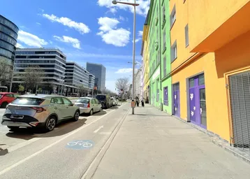 Blick auf eine belebte Stadtstraße mit parkenden Autos und farbenfrohen Gebäudefassaden unter blauem Himmel.