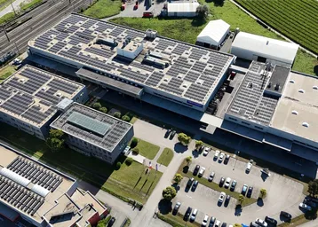 Luftaufnahme eines großen Industriekomplexes mit mehreren Gebäuden, Solaranlagen auf den Dächern und weitläufigen Parkplätzen.