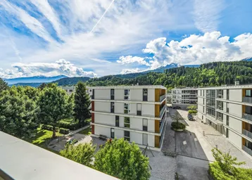 Blick auf moderne Wohngebäude mit grüner Umgebung und Bergen im Hintergrund unter blauem Himmel.