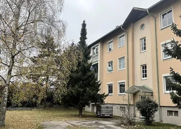Älteres Mehrfamilienhaus mit beiger Fassade, umgeben von Bäumen und einer Grünfläche.