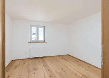 Helles Schlafzimmer mit Holzdielenboden, weiß gestrichenen Wänden und einem Fenster mit Blick ins Grüne.
