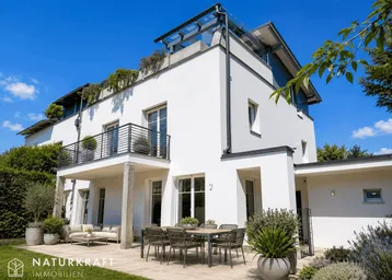 Moderne weiße Villa mit großer Terrasse, Gartenmöbeln und üppiger Bepflanzung unter blauem Himmel.