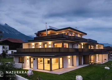 Modernes Mehrfamilienhaus bei Dämmerung mit beleuchteten Fenstern und Bergblick.