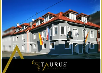 Graues Gebäude mit rotem Ziegeldach und mehreren Dachgauben an einer Straße.