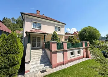 Einfamilienhaus mit Außentreppe, kleiner Terrasse und gepflegtem Vorgarten unter blauem Himmel.