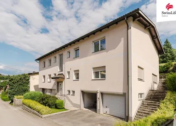 Mehrfamilienhaus mit Garagen und Außentreppe, umgeben von gepflegter Bepflanzung unter blauem Himmel.